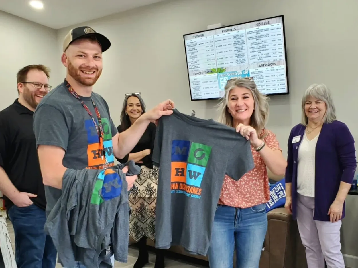 Group of people wearing matching gray t-shirts in dispensary interior with monitor displaying information
